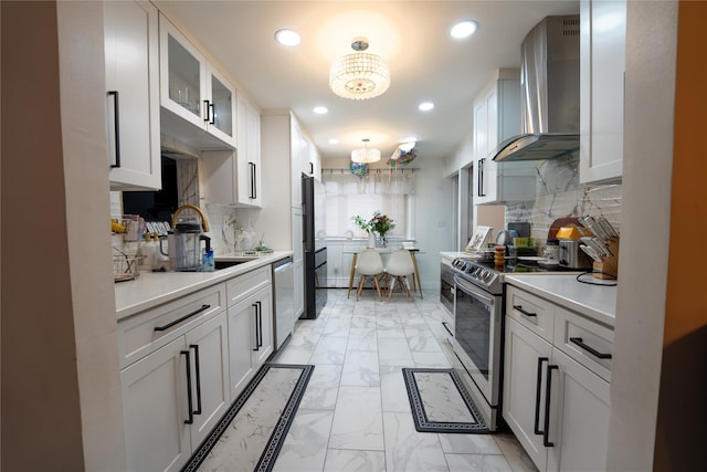 kitchen with wall chimney range hood, appliances with stainless steel finishes, light countertops, marble finish floor, and an inviting chandelier