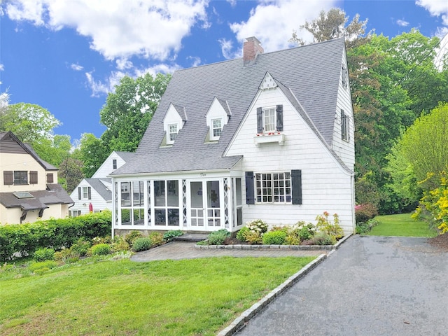 cape cod house with a sunroom, roof with shingles, a chimney, and a front yard