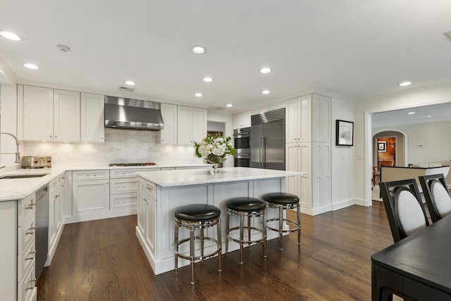 kitchen featuring wall chimney exhaust hood, appliances with stainless steel finishes, arched walkways, a center island, and a sink
