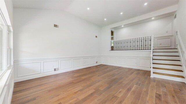 empty room featuring wood finished floors, a decorative wall, stairs, lofted ceiling, and wainscoting