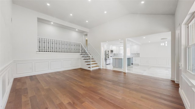 unfurnished living room with a decorative wall, stairs, light wood finished floors, wainscoting, and high vaulted ceiling