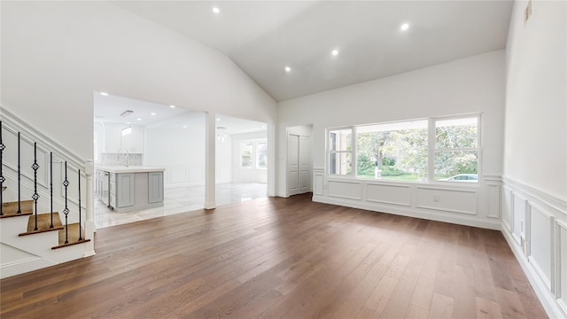 unfurnished living room with light wood-style flooring, a decorative wall, wainscoting, stairs, and high vaulted ceiling