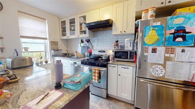 kitchen with stainless steel appliances, under cabinet range hood, backsplash, white cabinetry, and glass insert cabinets