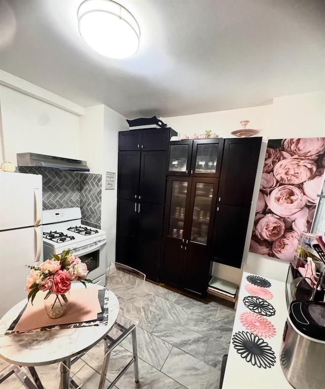 kitchen featuring glass insert cabinets, marble finish floor, dark cabinets, under cabinet range hood, and white appliances