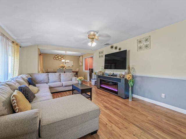 living room with ceiling fan with notable chandelier, light wood-type flooring, visible vents, baseboards, and a glass covered fireplace