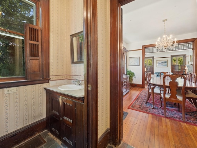 bathroom featuring an inviting chandelier, wood-type flooring, vanity, baseboards, and wallpapered walls