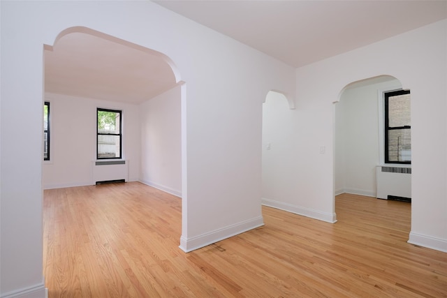 empty room featuring arched walkways, baseboards, radiator heating unit, and light wood-type flooring