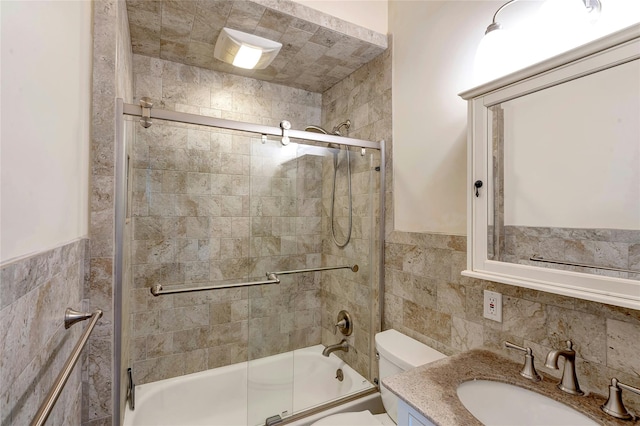 bathroom featuring tile walls, a wainscoted wall, vanity, toilet, and shower / bath combination with glass door