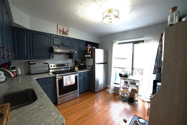 kitchen with light stone countertops, light wood-type flooring, under cabinet range hood, a sink, and stainless steel appliances