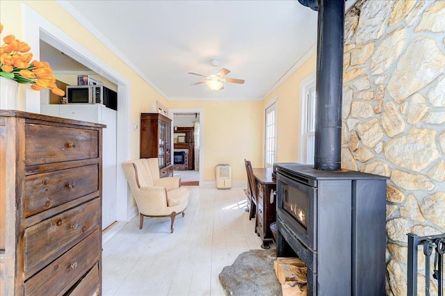 living room featuring ceiling fan, a wood stove, baseboards, light wood-type flooring, and ornamental molding