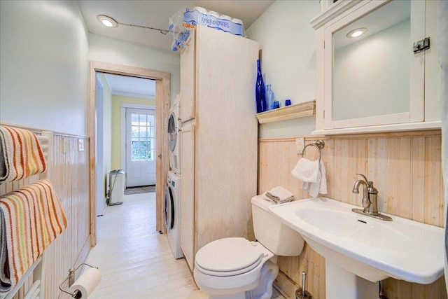 bathroom with wooden walls, wainscoting, a sink, toilet, and stacked washer and clothes dryer