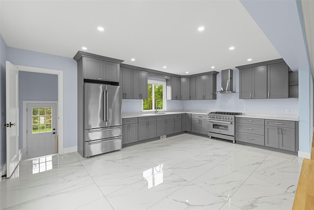 kitchen featuring baseboards, premium appliances, a sink, wall chimney range hood, and gray cabinets