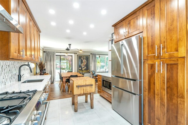 kitchen featuring stainless steel appliances, range hood, a sink, brown cabinetry, and tasteful backsplash