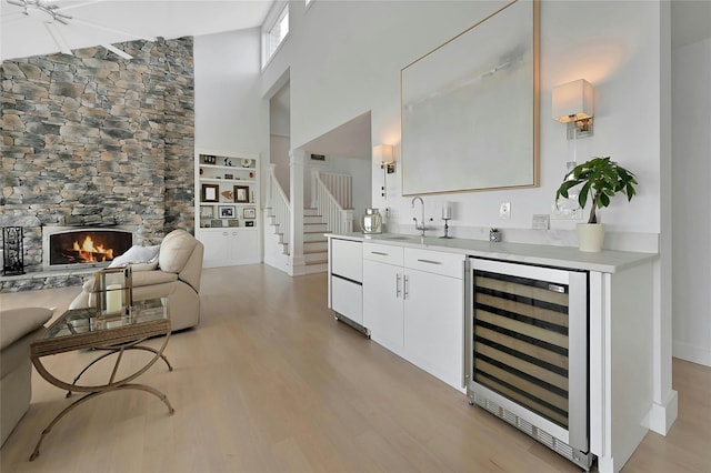 bar with a sink, a towering ceiling, light wood finished floors, beverage cooler, and a stone fireplace