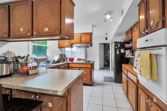 kitchen featuring a peninsula, brown cabinets, under cabinet range hood, light tile patterned floors, and appliances with stainless steel finishes