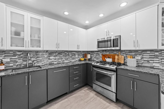 kitchen with stainless steel appliances, a sink, white cabinetry, dark stone counters, and glass insert cabinets