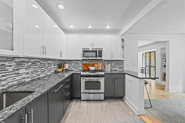 kitchen featuring stainless steel appliances, white cabinets, dark stone counters, a breakfast bar area, and glass insert cabinets