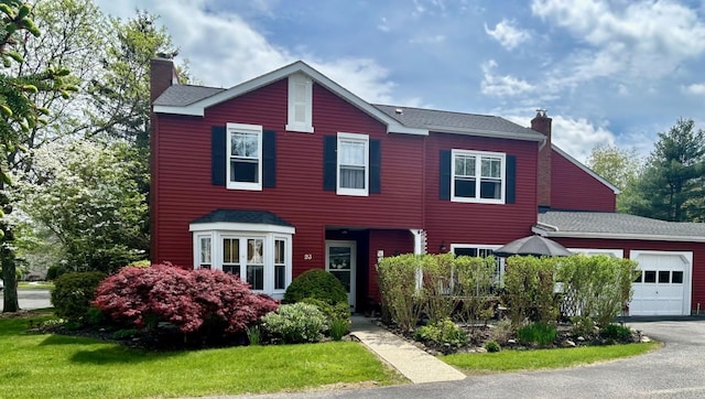 colonial-style house featuring roof with shingles, an attached garage, driveway, a front yard, and a chimney