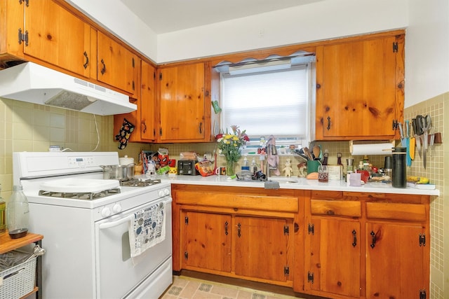 kitchen featuring tile walls, under cabinet range hood, a sink, brown cabinets, and white range with gas cooktop