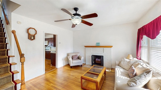 living room with stairway, ceiling fan, light wood-style floors, baseboards, and a brick fireplace
