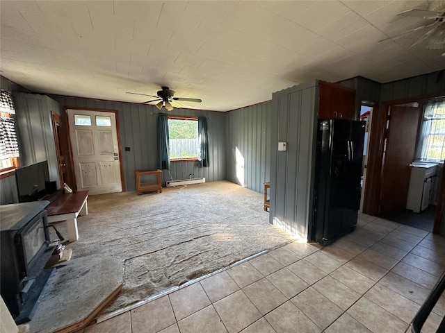 unfurnished living room featuring a ceiling fan, a wood stove, light tile patterned floors, and a baseboard radiator