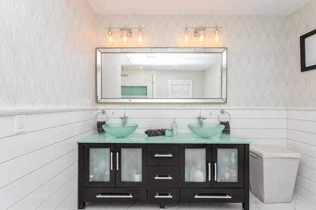 full bath with double vanity, wallpapered walls, a wainscoted wall, and a sink