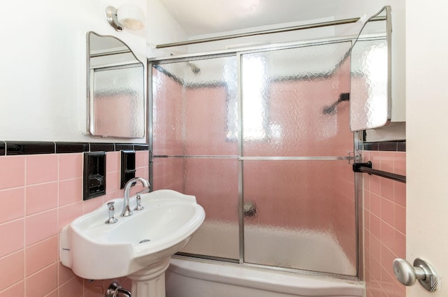 bathroom featuring tile walls, bath / shower combo with glass door, a sink, and wainscoting