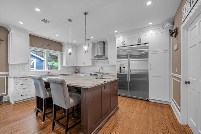 kitchen with stainless steel built in fridge, wall chimney range hood, an island with sink, tasteful backsplash, and white cabinets