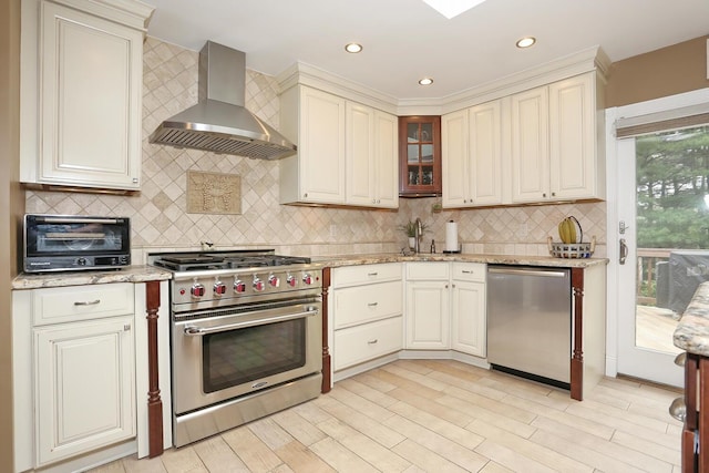 kitchen with stainless steel appliances, wall chimney exhaust hood, backsplash, light stone countertops, and cream cabinetry