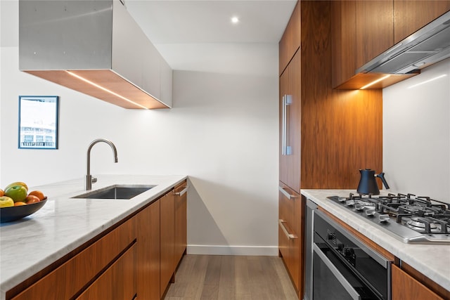 kitchen featuring a sink, brown cabinetry, stainless steel gas cooktop, wall chimney exhaust hood, and wall oven