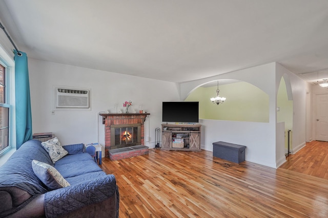 living area featuring a wall mounted air conditioner, light wood-type flooring, arched walkways, a brick fireplace, and a chandelier