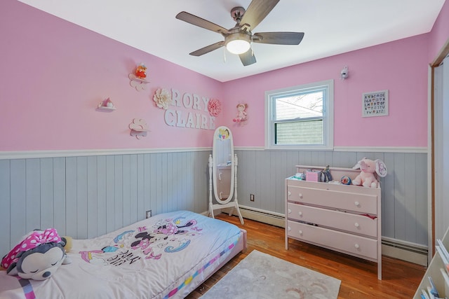 bedroom with wainscoting, wood finished floors, a baseboard radiator, and a ceiling fan