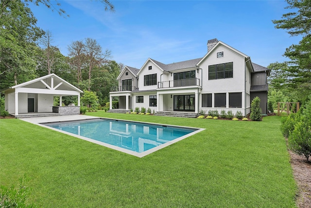 back of property with a balcony, a chimney, an outdoor pool, a lawn, and a patio
