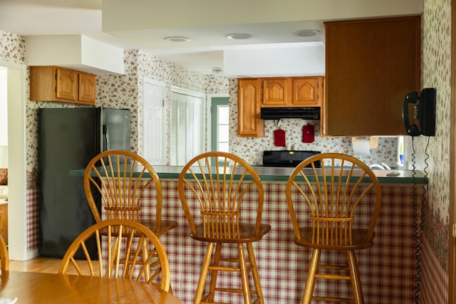 kitchen featuring freestanding refrigerator, wallpapered walls, under cabinet range hood, range, and brown cabinetry