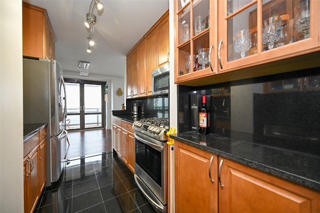 kitchen featuring appliances with stainless steel finishes, dark tile patterned flooring, brown cabinetry, glass insert cabinets, and dark stone counters