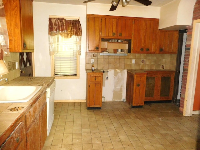 kitchen featuring tile countertops, ceiling fan, a sink, backsplash, and brown cabinets