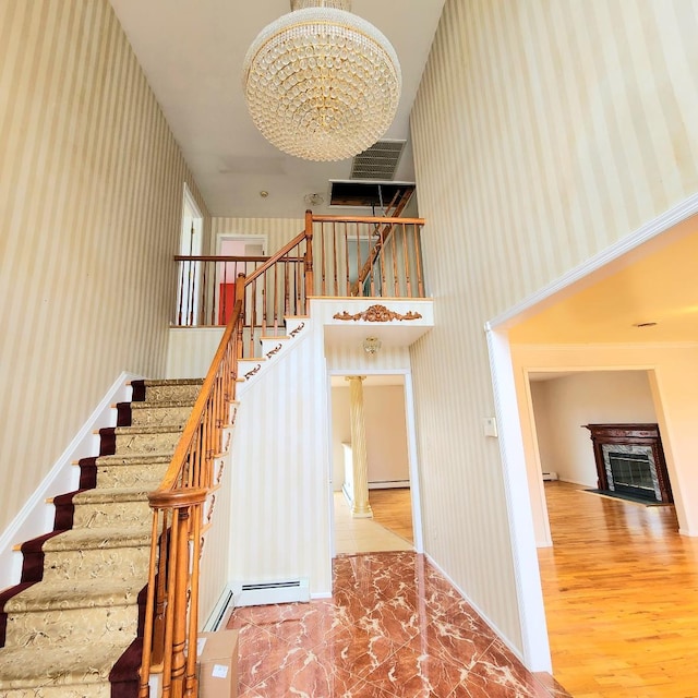 staircase featuring wallpapered walls, a chandelier, baseboard heating, a fireplace, and a towering ceiling