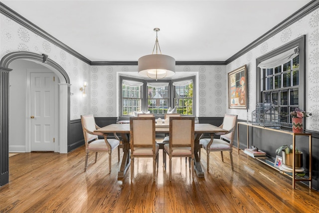 dining area with wallpapered walls, wood finished floors, ornamental molding, arched walkways, and a wainscoted wall