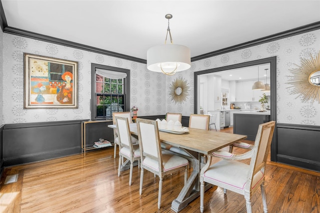 dining area featuring wallpapered walls, light wood finished floors, ornamental molding, and wainscoting