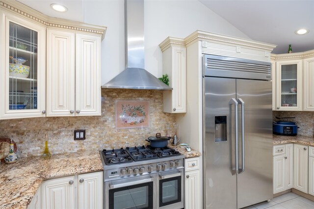 kitchen with premium appliances, wall chimney exhaust hood, cream cabinetry, backsplash, and light stone countertops