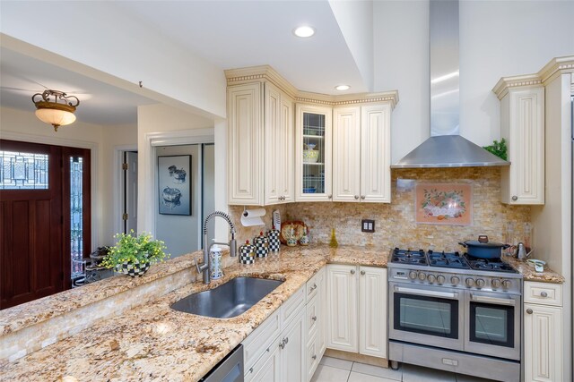 kitchen featuring double oven range, wall chimney range hood, a sink, cream cabinets, and light stone counters