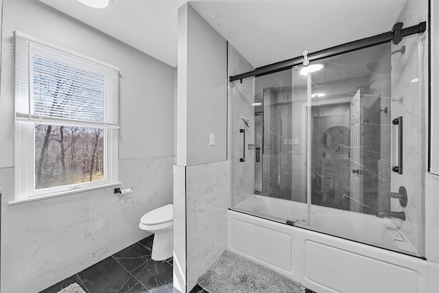 bathroom featuring marble look tile flooring, toilet, shower / bath combination with glass door, tile walls, and wainscoting