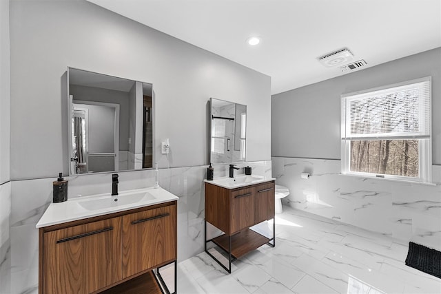 bathroom featuring tile walls, two vanities, toilet, a wainscoted wall, and marble look tile flooring
