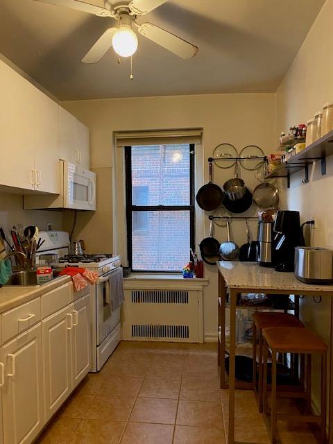 kitchen with radiator, white appliances, white cabinets, light tile patterned floors, and a ceiling fan
