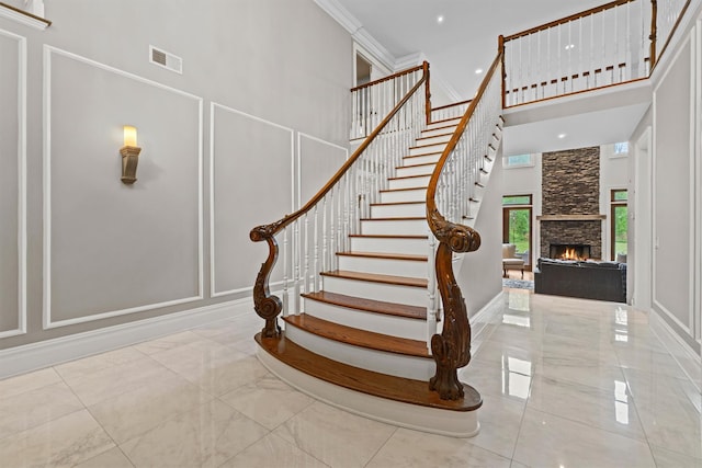 staircase featuring a decorative wall, a high ceiling, a stone fireplace, ornamental molding, and marble finish floors
