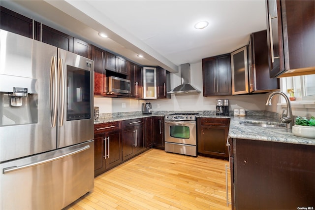 kitchen with stainless steel appliances, wall chimney exhaust hood, a sink, light wood-type flooring, and light stone countertops