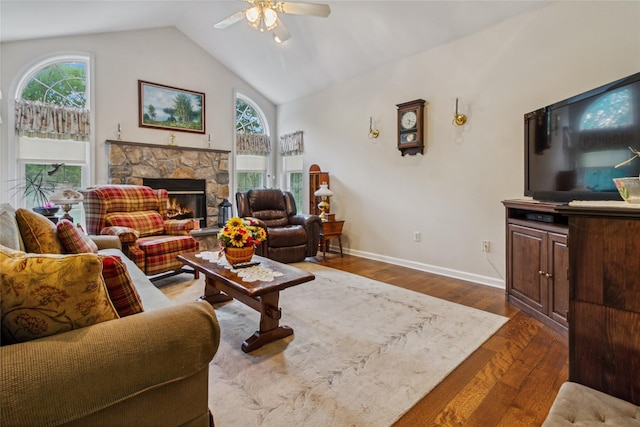 living room with ceiling fan, vaulted ceiling, dark wood-type flooring, baseboards, and a stone fireplace