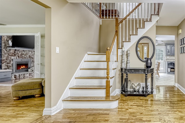 stairway with a fireplace, wood finished floors, baseboards, ceiling fan, and crown molding