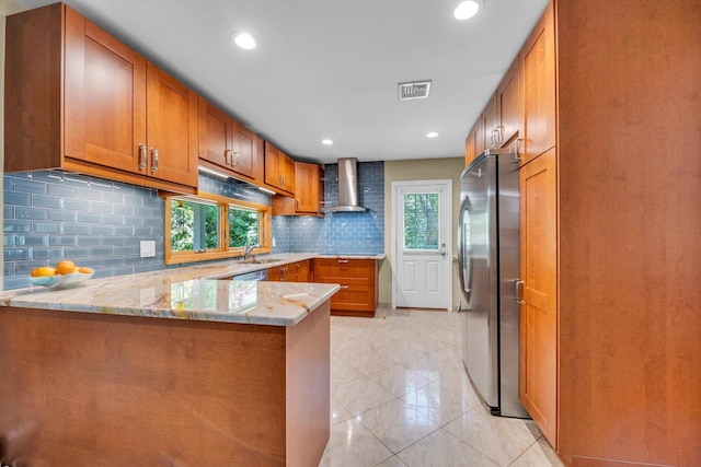 kitchen with freestanding refrigerator, wall chimney range hood, a peninsula, brown cabinetry, and recessed lighting