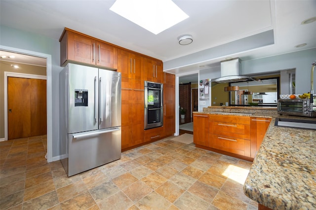 kitchen with stainless steel appliances, light stone finish flooring, wall chimney exhaust hood, brown cabinetry, and light stone countertops